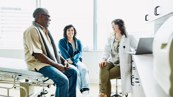 A female health care worker talks to an older man seated on a doctor’s table as a smiling young woman seated nearby looks on.