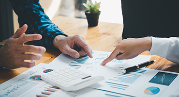 Two coworkers hands pointing out things on a series of graph charts on a desktop to illustrate reference-based pricing services. 