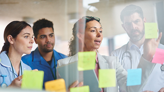 Group of healthcare workers working out a benefits administration problem with sticky notes on a clear glass wall.