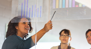 Three employees look up at graphs and information on a glass wall as one points out something, to illustrate benefits of self-funding.