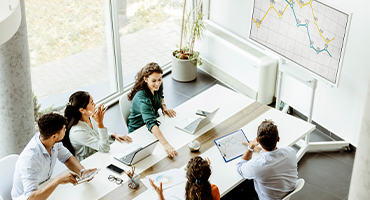 A group of employees sit around a long table with laptops open, smiling, talking and checking out a large graph on the wall about stop-loss insurance.