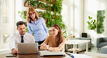 Two seated coworkers and one person standing all look at an open laptop to illustrate ease of benefits administration. 