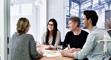 A group of coworkers viewed through a glass door as they sit around a table smiling and talking to illustrate benefits of having a self-funded plan.