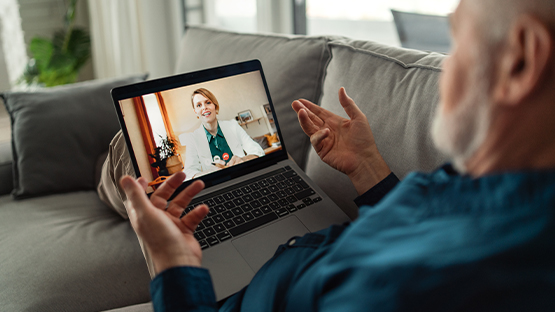 A bearded man gestures with his hands while chatting with a telehealth doctor on his open laptop to show benefits of a self-funded benefit plan. 