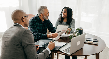 Two smiling black men and a smiling black woman sit at a small table strewn with an open laptop and insurance plan management services paperwork.