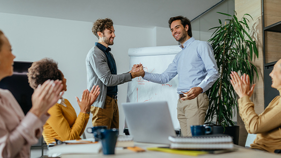 Two happy customers of health benefits administration stand in front of teammates and clasp hands while seated teammates applaud.